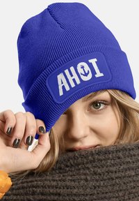 Blue knitted beanie featuring a white patch with "AHOI" and anchor design. Textured fabric with a snug fit, rolled hem for style.