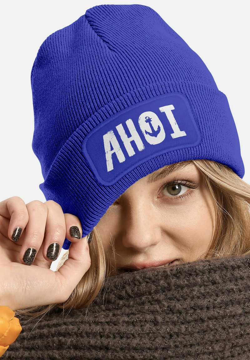 Blue knitted beanie featuring a white patch with "AHOI" and anchor design. Textured fabric with a snug fit, rolled hem for style.