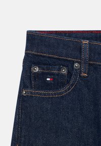 Σκούρο μπλε τζιν παντελόνι με μπροστινή τσέπη που διαθέτει μεταλλικά αξεσουάρ και μικρή ετικέτα λογότυπου σε κόκκινο, άσπρο και μπλε χρώμα. Ορατή αντίθεση στη ραφή.