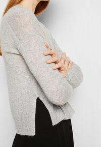 Hellgrauer Strickpullover mit langen Ärmeln, gerippten Bündchen und einem dezenten seitlichen Schlitz. Weiche Textur mit etwas lockerer Passform.