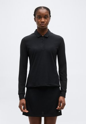 Ženska, ki nosi črno srajco z dolgimi rokavi in ovratnikom ter črno krilo, stoji pred enostavnim svetlim ozadjem.