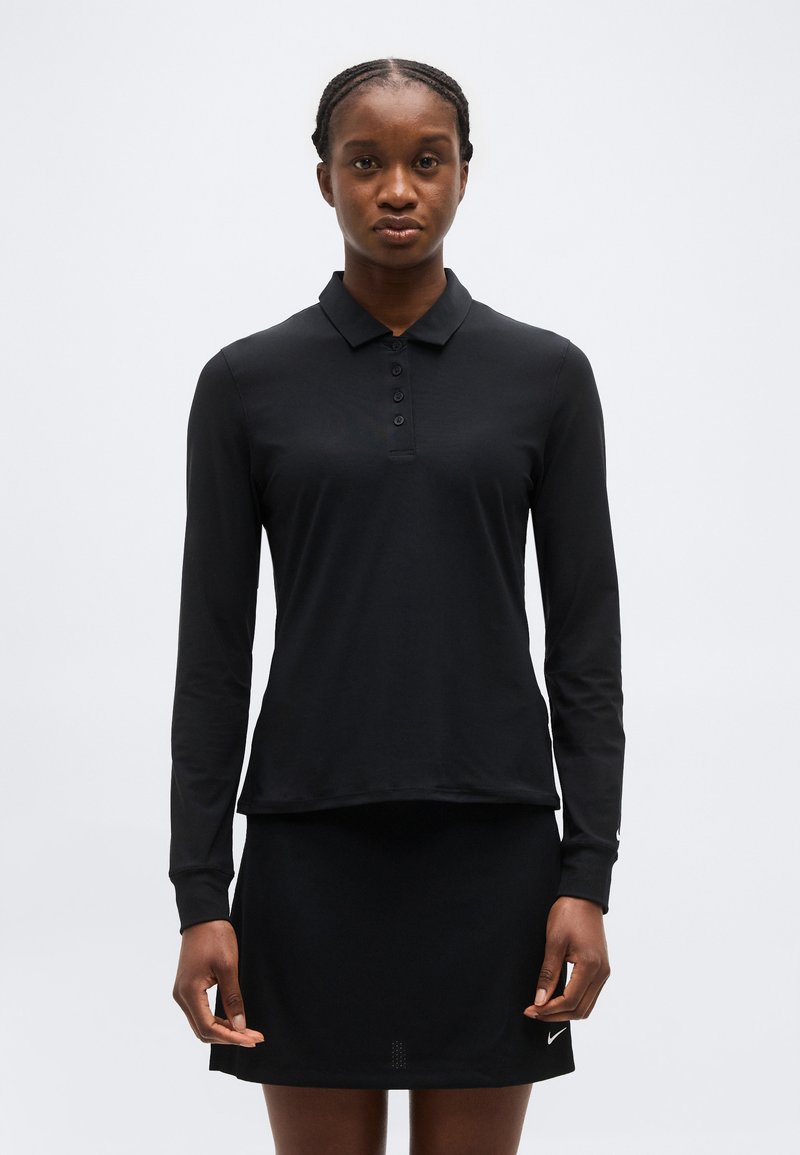 Γυναίκα που φορά μια μαύρη μπλούζα με μακριά μανίκια και γιακά και μια μαύρη φούστα, στέκεται μπροστά σε απλό ανοιχτό φόντο.