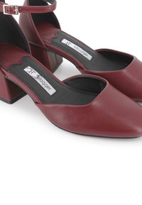 Zapatos de cuero burdeos con tacón bajo y ancho, con correas en el tobillo, punta afilada y suelas interiores negras etiquetadas como "blogger" sobre un fondo blanco.