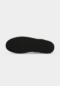 Semelle de chaussure en caoutchouc noir avec un motif de bande de roulement triangulaire répétitif sur un fond clair neutre.