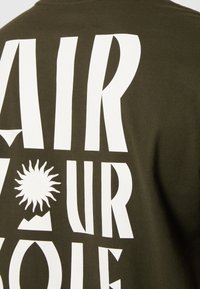 Olivově zelené bavlněné tričko s velkou bílou typografií a grafikou slunce a hor. Design zdůrazňuje výrazný text a minimální detaily.