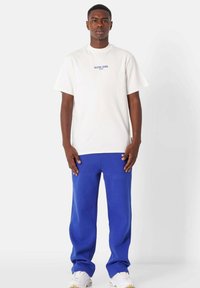 Hvid bomulds t-shirt med blå tekst, parret med blå shorts med et lille logo. Modellen står imod en lys grå baggrund.
