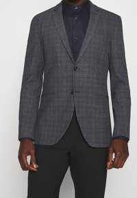 Blazer v tmavé šedé houndstooth vzoru, s dvouknoflíkovým zapínáním, klopami a vystuženými rameny. Spárováno s tmavou košilí na knoflíky.