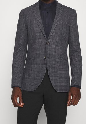 Blazer in donkergrijze houndstoothprint, met een dubbele knoopsluiting, ingesneden revers en gestructureerde schouders. Gecombineerd met een donker overhemd.