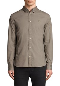 Mężczyzna w szarej koszuli z długim rękawem i zapięciem na guziki, z małym haftowanym logo na lewej piersi, w połączeniu z czarnymi spodniami.