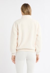 Pullover in pile crema chiaro, con collo alto, maniche a palloncino e vita aderente. Tessuto morbido; dettagli minimi; colore uniforme.