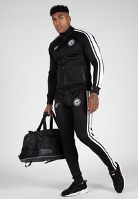 Zwart sportief trainingspak met witte strepen, ritsjack, bijpassende broek en een zwarte sporttas, aangevuld met zwarte sneakers.