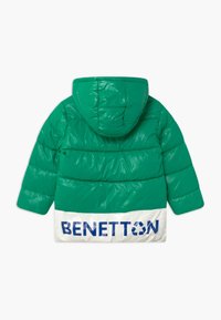 Grön pufferjacka med huva, med en glänsande yta och ribbad textur, samt en vit nederdel med "BENETTON" tryckt i blått.