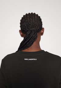 Svart bomullströja med rund halsringning, med "KARL LAGERFELD" broderat i vitt på övre delen av ryggen, som visar flätad frisyr.