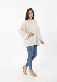 Maglione oversize beige con collo a costine e maniche a palloncino, abbinato a jeans in denim blu aderenti e sandali con tacco quadrato trasparente.