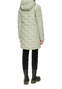 Helle grüne Steppjacke mit Kapuze, die eine gesteppte Textur aufweist. Kombiniert mit schwarzen Hosen und Knöchelstiefeln, was ein minimalistisches Design betont.