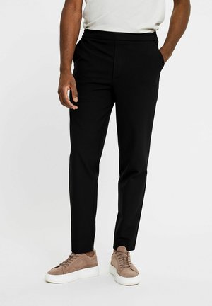 Man draagt zwarte slim-fit broek en beige suède sneakers, staand met één hand in de zak tegen een witte achtergrond.