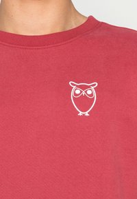 Red sweatshirt made of cotton, featuring a white owl graphic on the left chest. Round neckline with ribbed trim. Smooth texture.