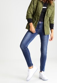 Olivově zelená pracovní bunda přes černý top, spárovaná s modrými skinny džínami a bílými teniskami, která ukazuje ležérní, vrstvený vzhled.