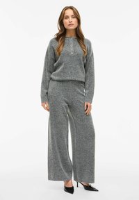 Szary sweter dzianinowy z wysoką szyją i pięcioma guzikami, zestawiony z szerokimi spodniami w tym samym kolorze. Tkanina ma miękką teksturę.