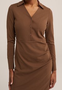 Brun långärmad klänning med en urtagning krage, knappstängning och draperad tygdetalj, tillverkad av ett mjukt, stretchigt material.