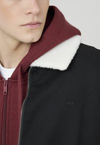 Veste à capuche avec un col en polaire bordeaux et une fermeture éclair à l'avant. Le corps est noir avec une texture côtelée et un logo en relief.