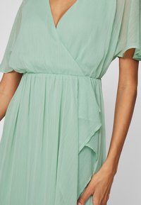 Lichtgroene geplooide jurk met V-hals en doorschijnende korte mouwen, gedragen door een persoon met een zichtbaar arm aan de zijkant.