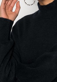 Pullover nero lavorato a maglia con collo alto, maniche larghe e una texture morbida. Include una mano sollevata e un grande orecchino circolare visibile.