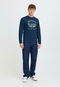 Marineblaues Langarm-T-Shirt mit "BLEND" Logo in Grün und Weiß, kombiniert mit dunkelblauen Jeans und weißen Sneakers, vor einem hellen Hintergrund stehend.