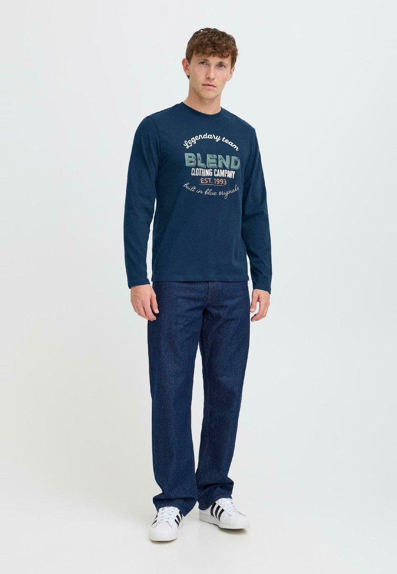 Marineblaues Langarm-T-Shirt mit "BLEND" Logo in Grün und Weiß, kombiniert mit dunkelblauen Jeans und weißen Sneakers, vor einem hellen Hintergrund stehend.