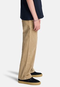 Personne portant un pantalon en velours côtelé beige, des baskets noires avec semelles en caoutchouc, une chemise noire et un bracelet tressé, debout devant un fond blanc.