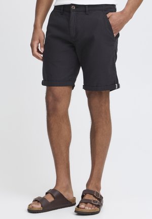 Mann trägt schwarze gekrempelte Shorts und braune Sandalen mit doppeltem Riemen, steht mit einer Hand in der Tasche vor weißem Hintergrund.