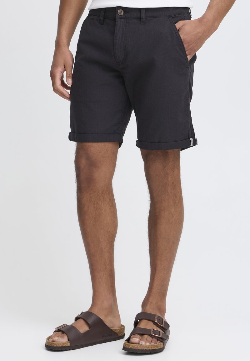 Homme portant un short noir retroussé et des sandales marron à double brides, debout avec une main dans la poche sur fond blanc.