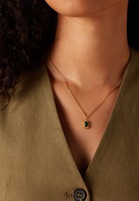 Gouden ketting met een smaragd geslepen groene edelsteen in een gestructureerd gouden frame, gedragen op een diepe V-hals olijfgroene kleding.