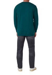 Teal sweatshirt met lange mouwen, geribbelde zoom en ronde hals, gecombineerd met donkergrijze jeans en witte sneakers, van achteraf bekeken.
