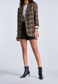 Brązowa marynarka w kratę z pojedynczymi guzikami, klapami z wycięciem i podwiniętymi rękawami, noszona na koszulce z nadrukiem i czarnych szortach, zestawiona z czarnymi botkami za kostkę.