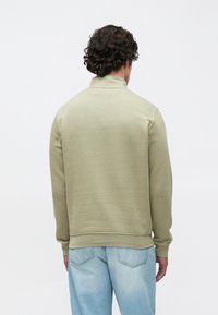 Roheline pusa kõrge kaelusega, pehme tekstuuriga ja soonikkoes varrukate äärtega. Kandmisega koos hele siniste teksadega, nähtav tagantpoolt.