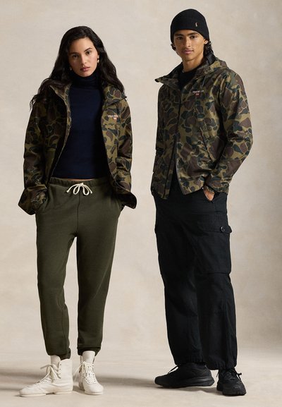 Casacos camuflados em verde e castanho, combinados com calças cargo pretas e calças de treino verdes-oliva. Os acessórios incluem um gorro preto e ténis brancos.