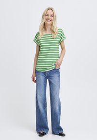 Mosolygó nő, hosszú szőke hajjal, zöld-fehér csíkos ingben, bő szárú kék farmerben és fekete cipőben, zsebébe dugott kézzel áll.