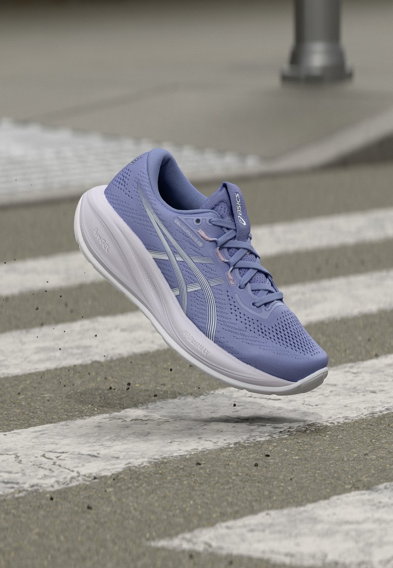 Μπλε αθλητικό παπούτσι με λευκή σόλα, αιωρούμενο πάνω από διάβαση πεζών σε ασφάλτινο δρόμο, που δείχνει κίνηση και αστικό υπαίθριο περιβάλλον.