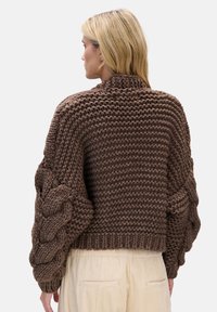 Brązowy sweter z dzianiny o wysokim kołnierzu i grubych rękawach w wzór warkocza. Teksturowana przędza i krótki dół. Noszony z jasnymi spodniami.