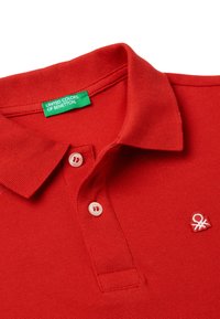 Rotes Poloshirt aus strukturiertem Stoff, mit klassischem Kragen, zwei weißen Knöpfen und einem gestickten Logo auf der Brust. Grünes Markenschild sichtbar.