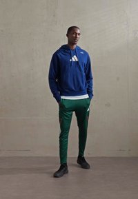 Egy férfi áll egy egyszínű falnak támaszkodva, kék Adidas kapucnis pulcsiban, zöld Adidas melegítőnadrágban és fekete sportcipőben.