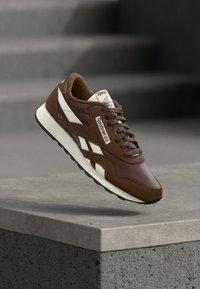 Smeđi i bijeli Reebok tenisica suspendirana iznad betonskog zida s zamagljenom sivoj pozadinom.