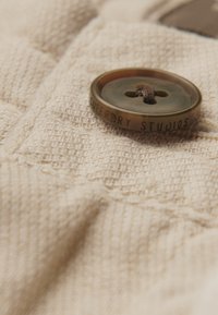 Tejido texturizado beige con un primer plano de un botón redondo en tonos tierra con "SUPERDRY STUDIOS" grabado.
