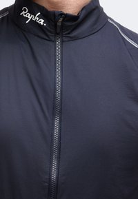Záber zblízka na osobu v námornícky modrej bunde na zips s logom „Rapha“ na golieri a reflexnými pruhmi na ramene.
