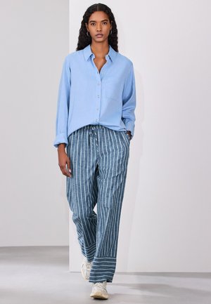 Femme aux longs cheveux bouclés porte une chemise bleu clair boutonnée, un pantalon bleu rayé ample et des baskets blanches, marchant sur un fond neutre.