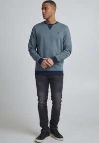 Lyseblå crewneck sweatshirt med navy detaljer på manchetterne og kanten, parret med mørke jeans og sorte sneakers, præsenteret på en neutral baggrund.