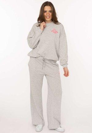 Junge Frau lächelt, trägt hellgraue Jogginghose und Sweatshirt mit dem Schriftzug „Feel Good Club“ und weiße Sneaker, steht vor weißem Hintergrund.