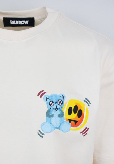 Camiseta de algodón color crema que presenta un gráfico de un oso azul con X en los ojos y una cara sonriente amarilla con tres dientes negros.