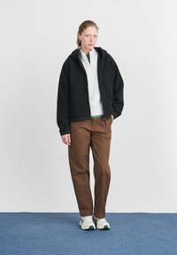 Black fleece zip jacket over a gray sweatshirt and green shirt, paired with brown trousers and white sneakers with green accents.